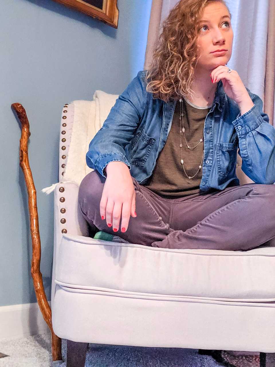 Hannah Donor wearing grey jeans, a green tshirt, and a denim button-down sitting cross-legged on a modern white chair thoughtfully looking upwards