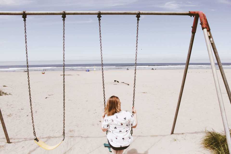 Mom sitting on a swingset on the beach in Seaside, Oregon while her children play