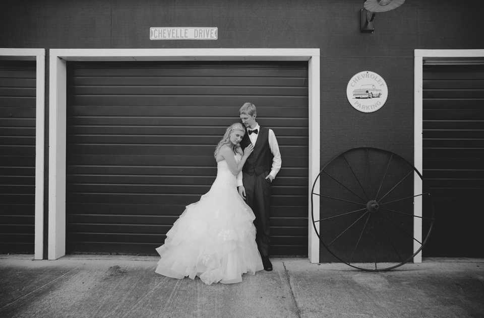 Black and white wedding photo of bride wearing white dropped waist ballgown and groom in vest and bowtie standing in front of a rustic garage. The bride is leaning on the groom's shoulder with her hand on his chest. There is an old rusty wagon wheel next to them and a sign above that reads, "Chevelle Drive."