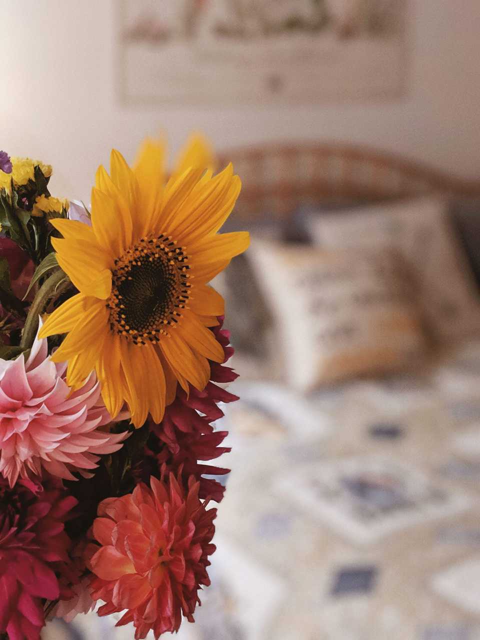 Flower bouquet with sunflower in foreground and rustic neutral toned bed with handmade quilt blurred in background