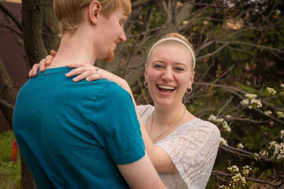 Romantic outdoor photo of husband Josiah in blue tshirt with arms on waist of wife, Hannah, in white lace shirt laughing with her hands on hubby's shoulders