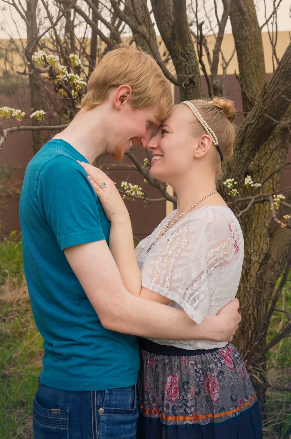 Happy husband and wife embrace nose-to-nose in this romantic outdoor setting. Husband Josiah is wearing jeans and a blue vneck tee and wife Hannah is wearing a white lace top, peasant skirt, and pearl headband